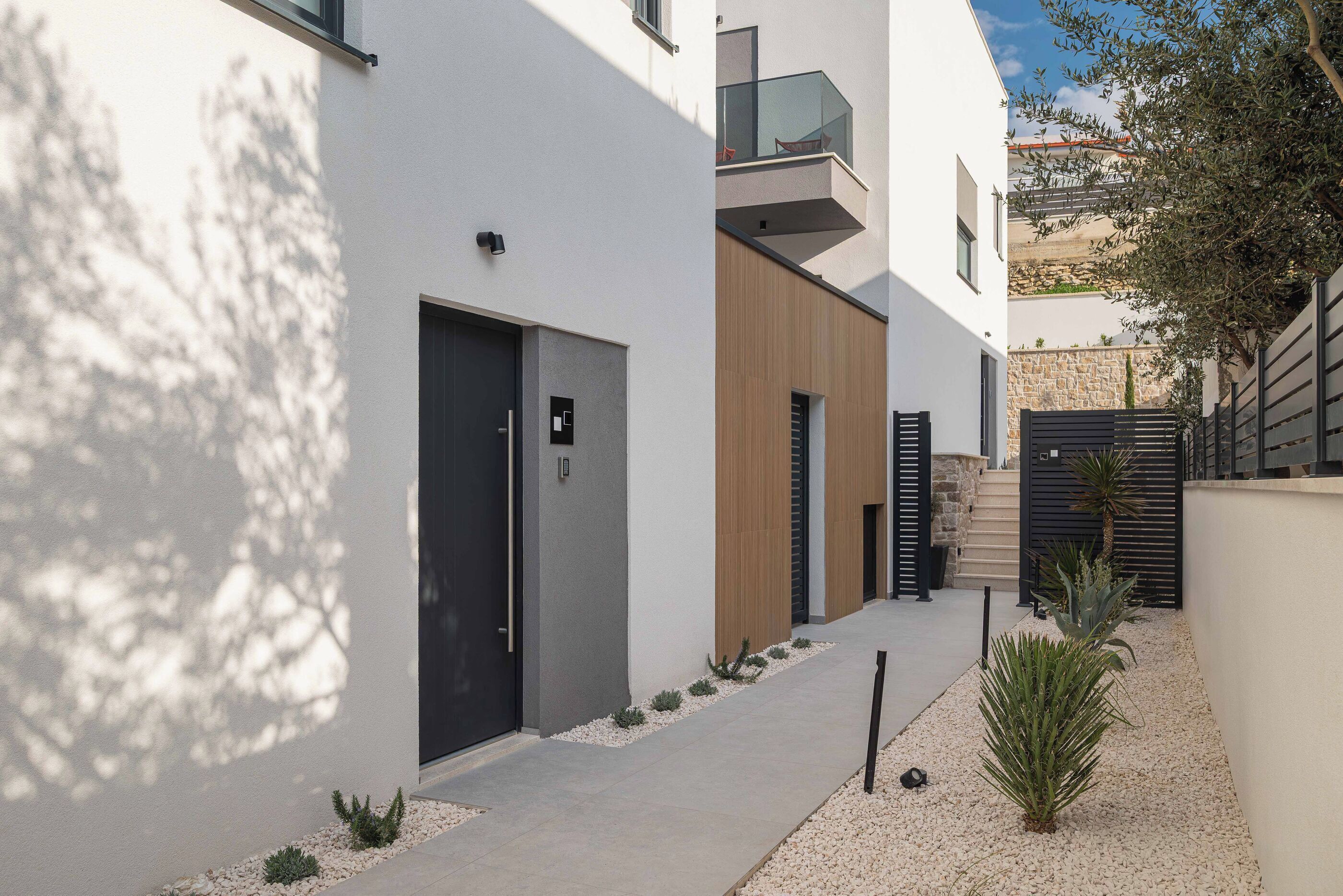 Un bâtiment moderne à la façade sobre, doté d’une porte noire et d’un petit jardin agrémenté de plantes et de gravier.