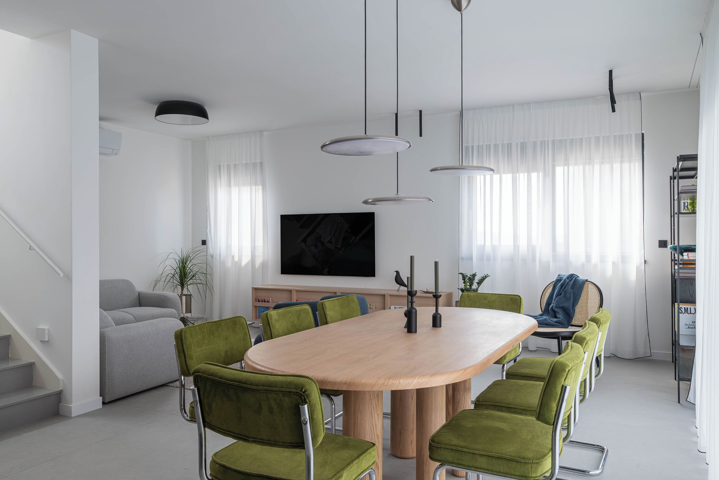 Ein moderner Essbereich mit einem runden Tisch, grünen Polsterstühlen, Pendelleuchten und einem großen Flachbildfernseher an der Wand.