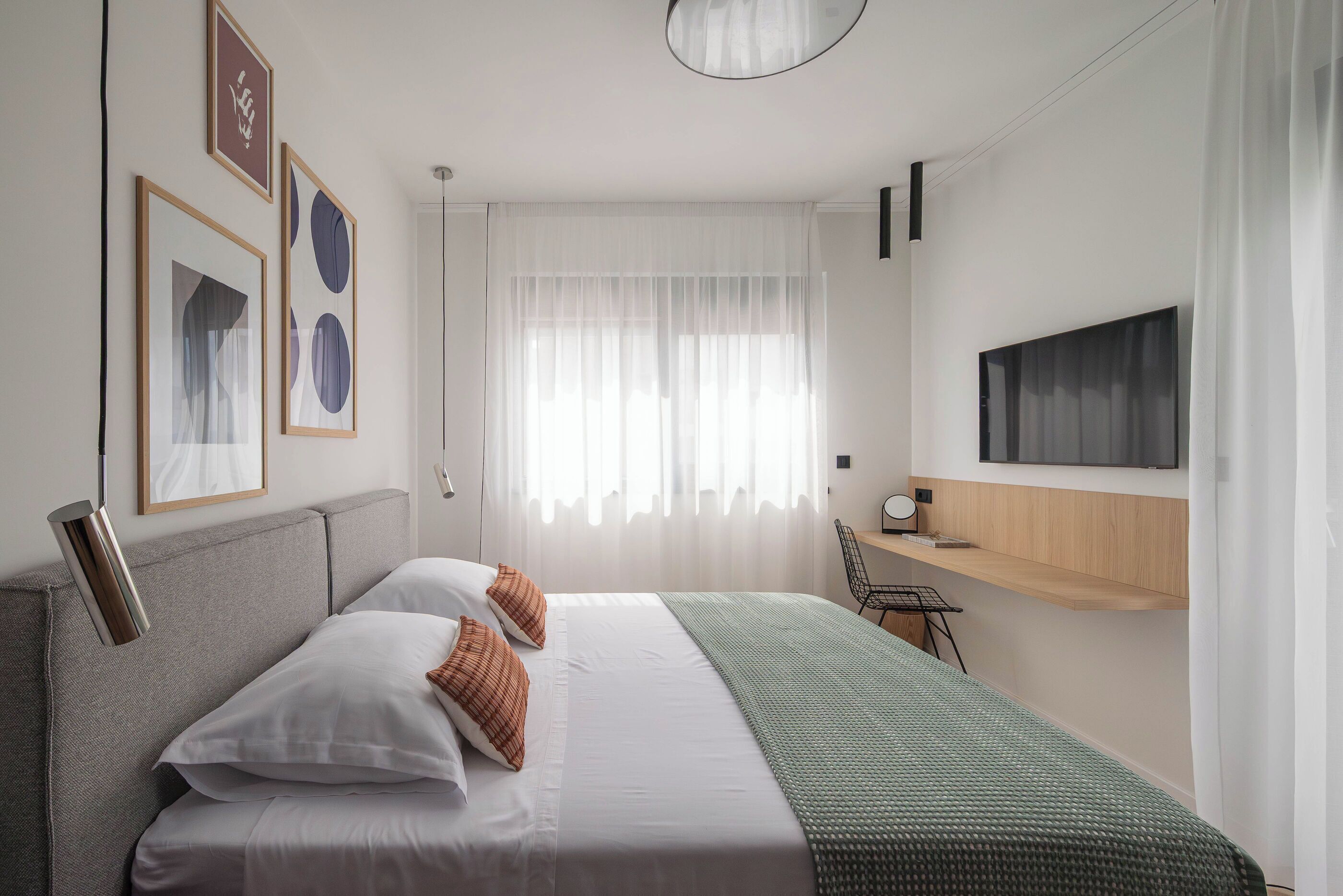 A modern hotel room with a bed, a desk, a TV, and a window with sheer curtains.