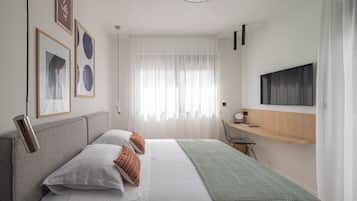 A modern hotel room with a bed, a desk, a TV, and a window with sheer curtains.