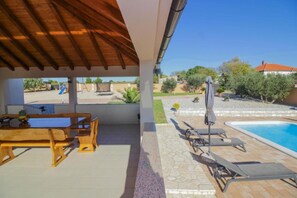 A covered patio with a wooden table and chairs, a poolside area with lounge chairs, and a view of a swimming pool and surrounding landscape. - Villa Zrilic With Private Pool (Benkovac)