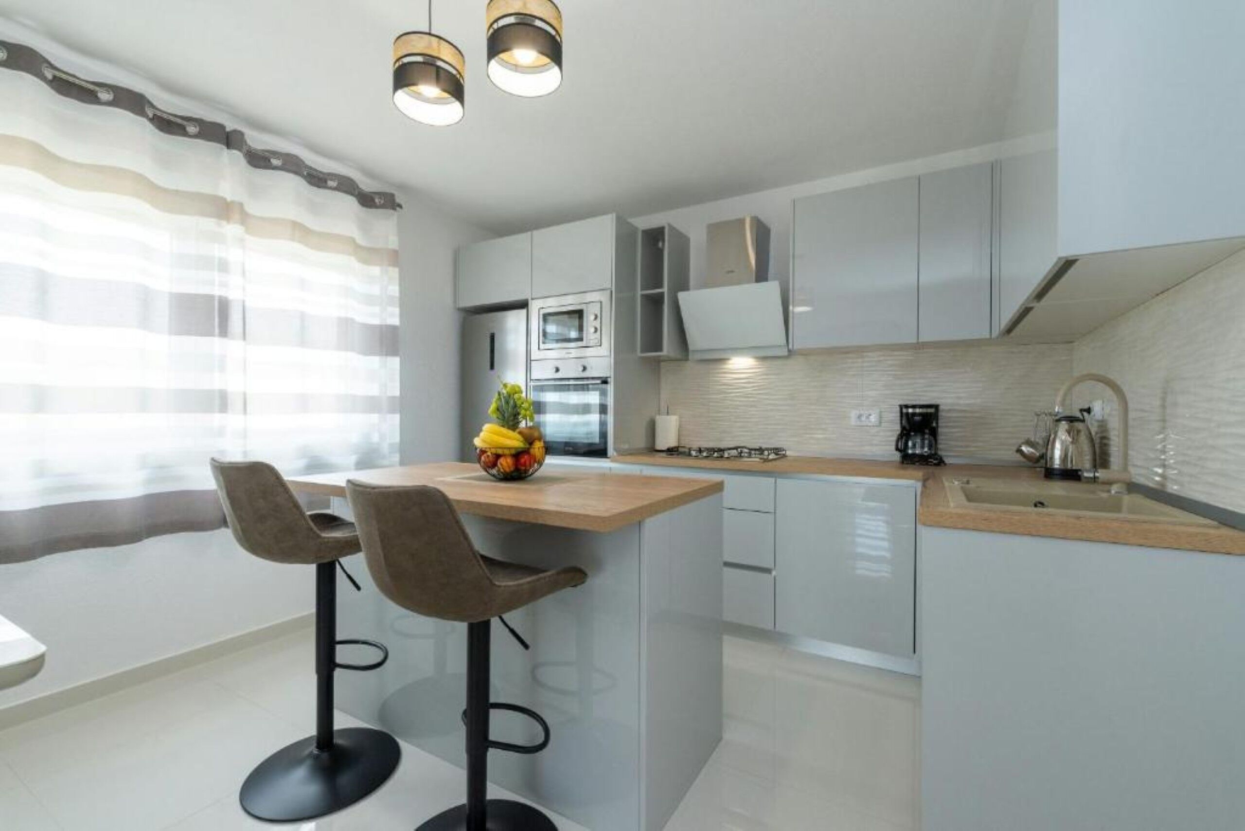 A modern kitchen with a central island, bar stools, a fruit bowl, and a coffee maker.