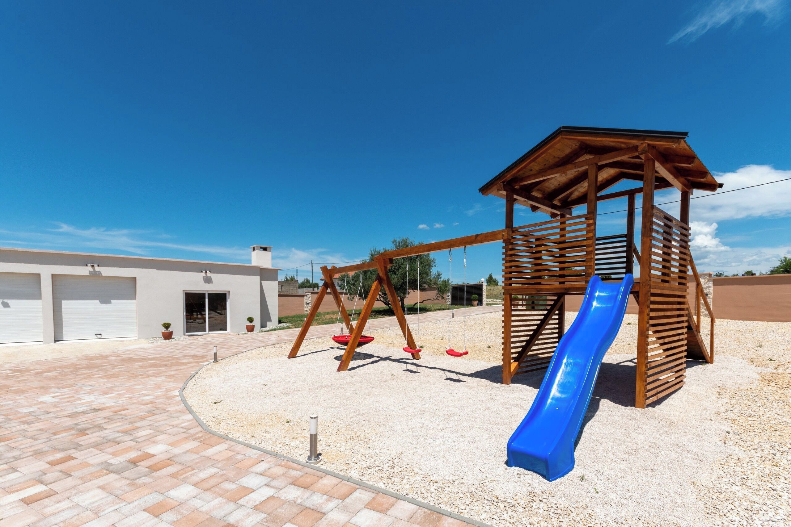 A playground with a wooden slide and swings, a covered play structure, and a building with garage doors in the background.