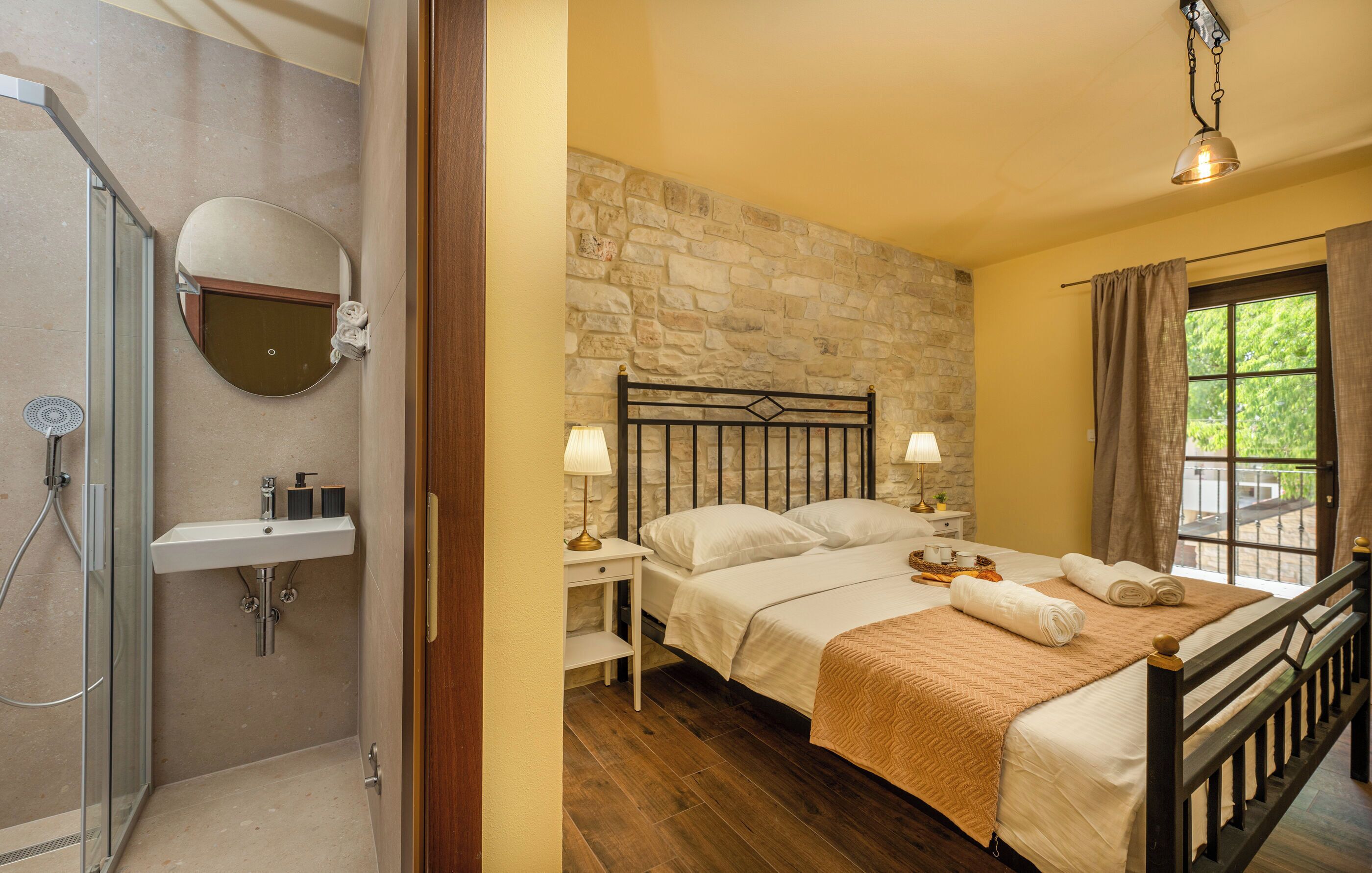 Un dormitorio con pared de piedra, cama con ropa blanca, mesita de noche con lámpara y ventana con cortinas.