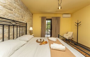 Ein Hotelzimmer mit Steinwand, einem Bett mit weißen Leinentüchern, einer Ablage mit Brot und Kaffee, einem Stuhl mit Handtuch und einer Klimaanlage.
