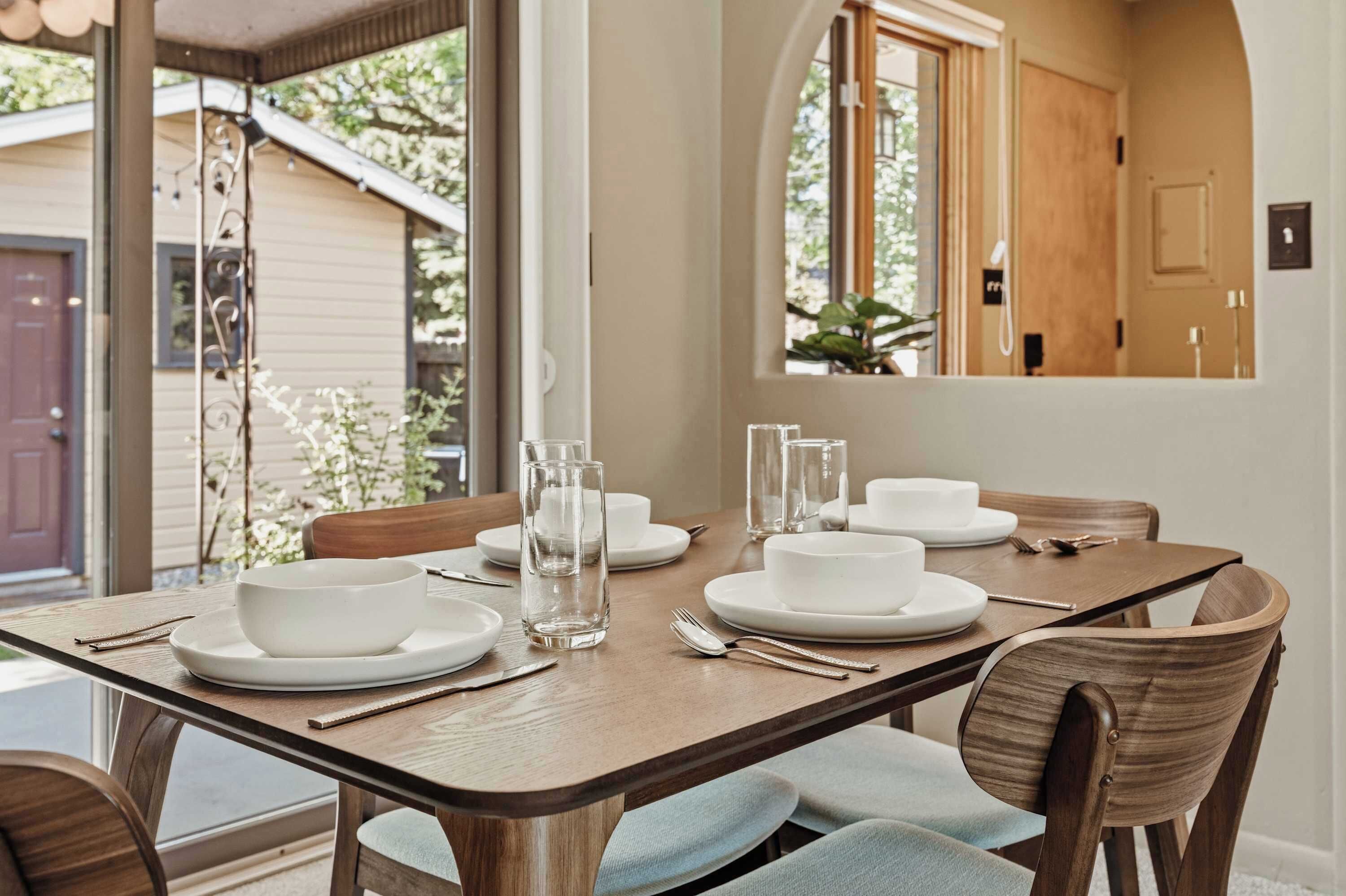 Mesa lista con platos, tazones, vasos y cubiertos blancos, con vista a una casa y follaje exterior.