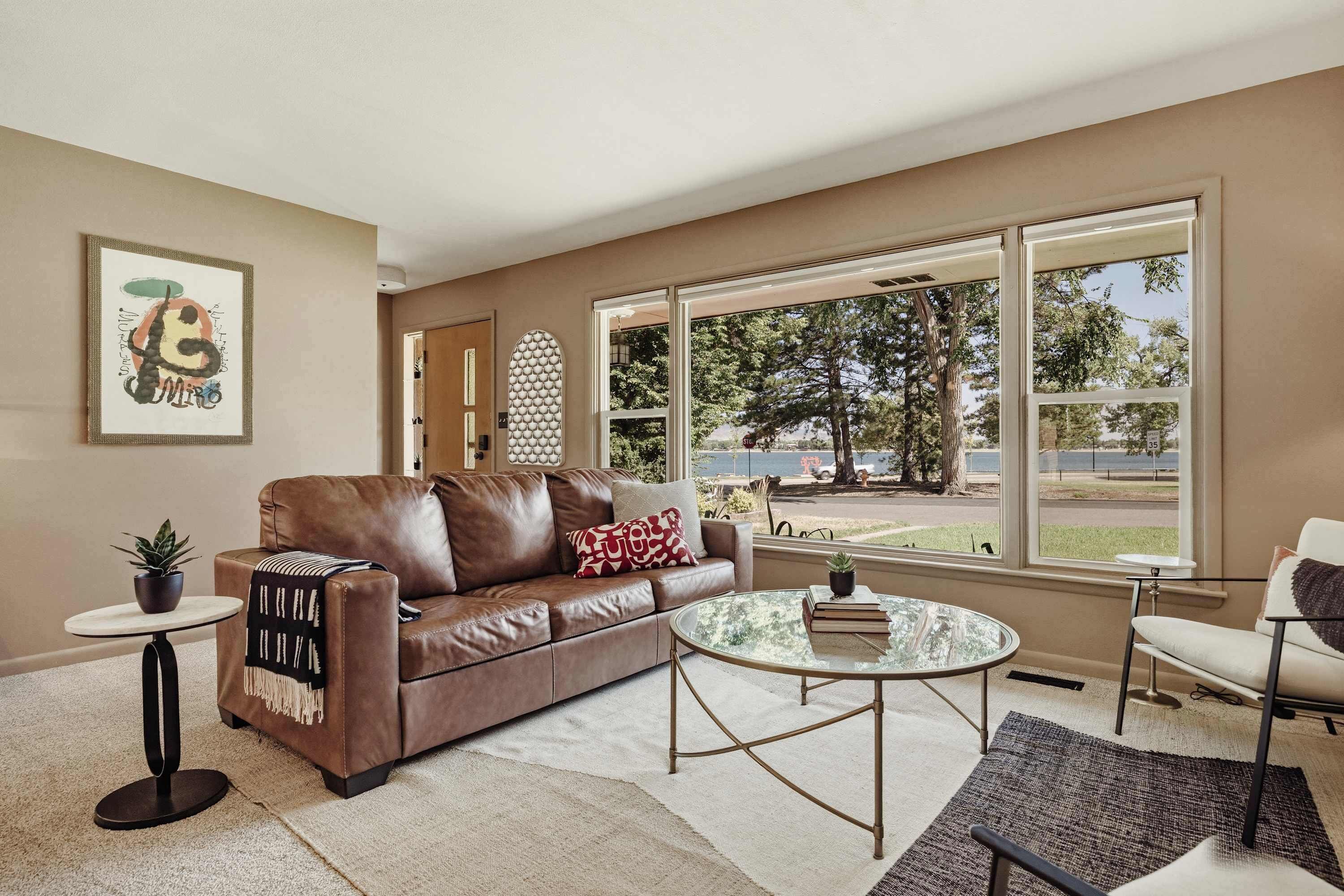 A living room with a brown leather sofa, a round glass coffee table, and a view of a park and lake through large windows.