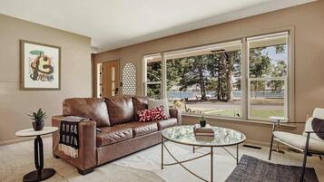 A living room with a brown leather sofa, a round glass coffee table, and a view of a park and lake through large windows.
