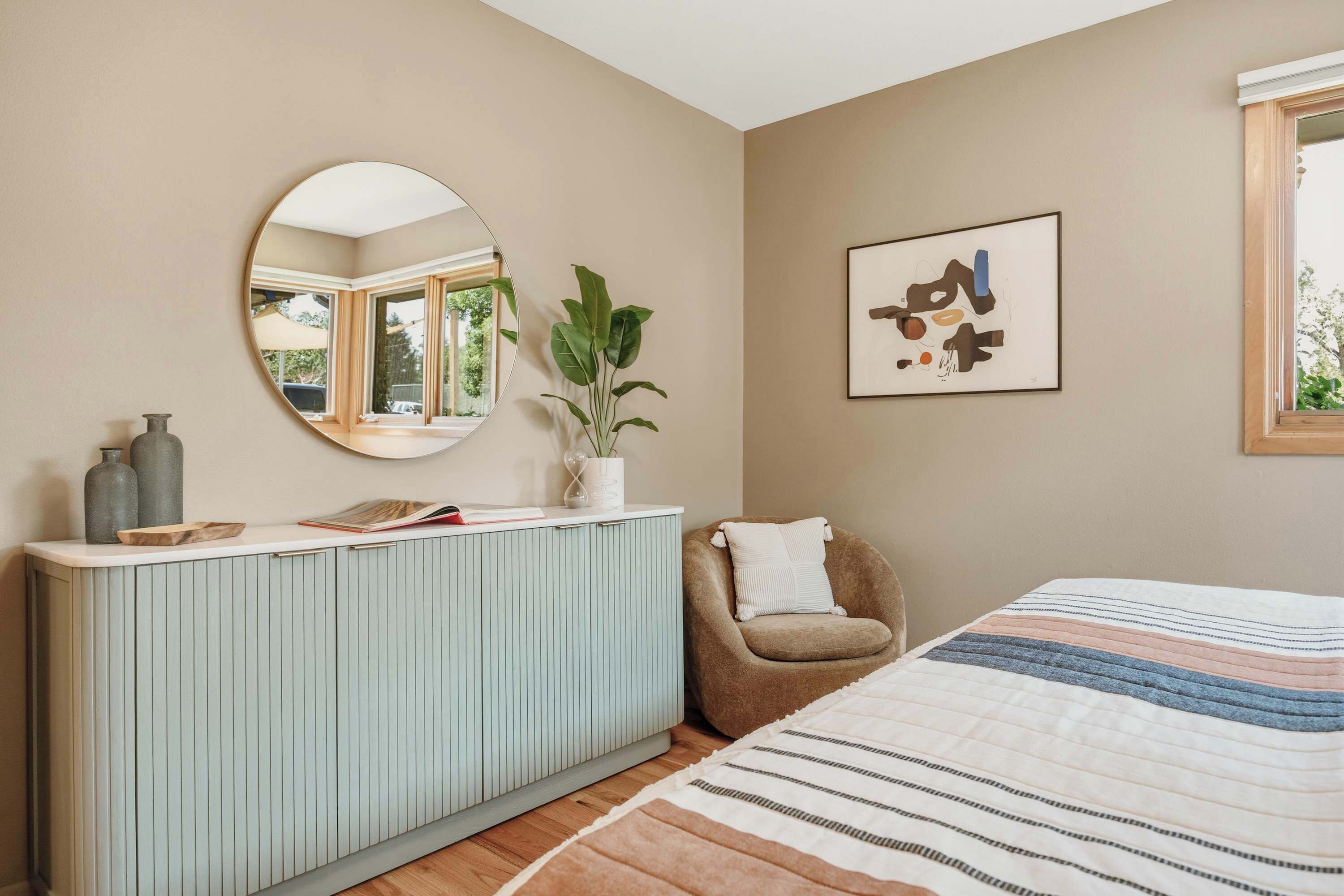 A bedroom with a bed, a chair, a bedside table, a mirror, a plant, and a framed artwork on the wall.