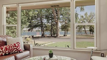 A room with a leather couch, a glass coffee table with books, and a view of a park and lake through large windows.