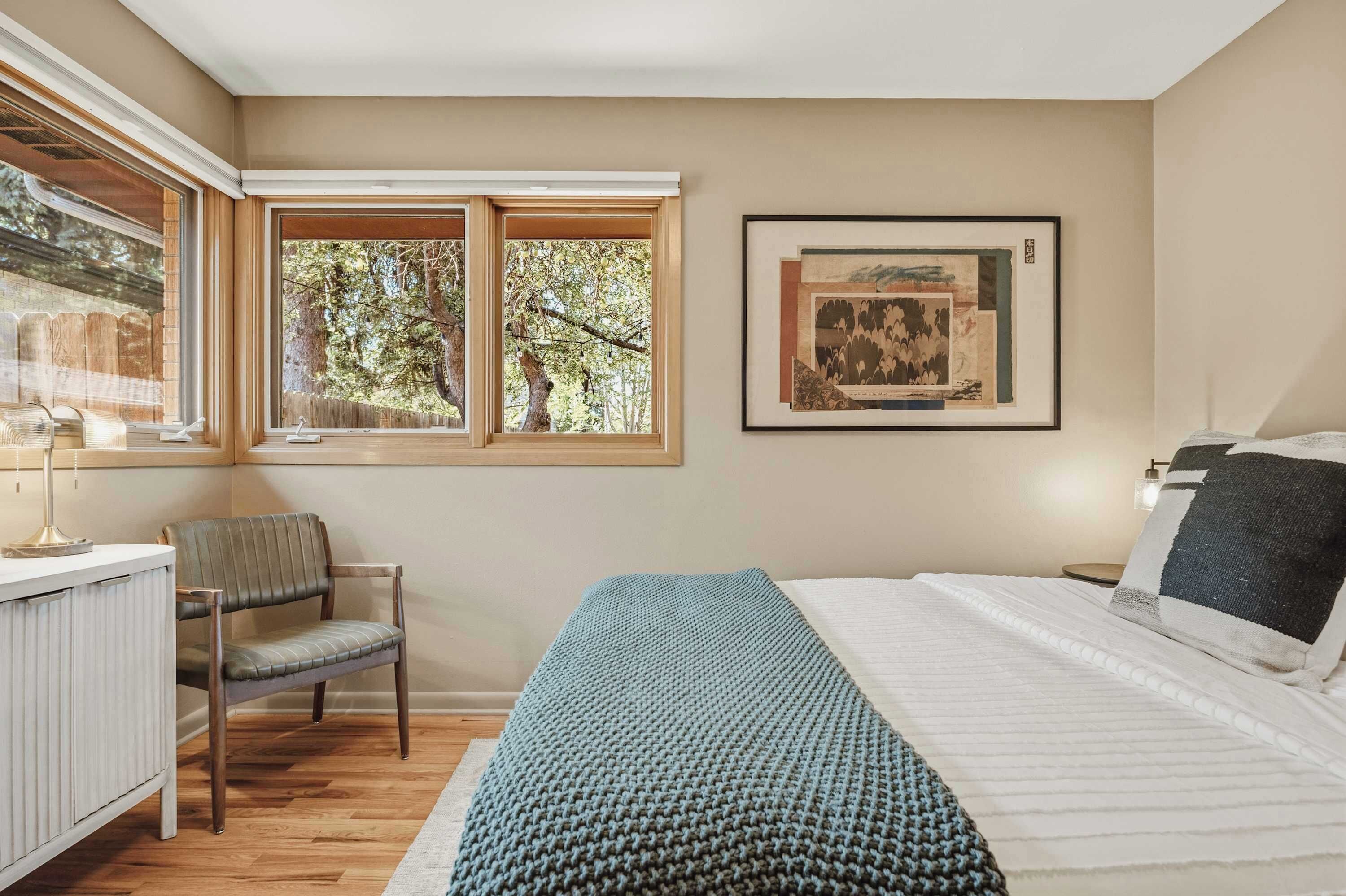 A bedroom with a bed, a desk, a chair, a window with a view of trees, and a framed artwork on the wall.