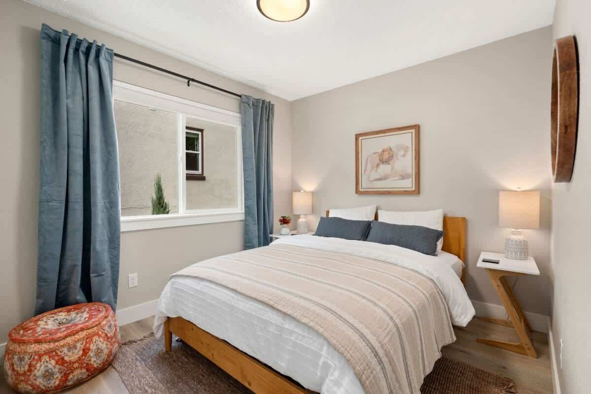 Een slaapkamer met een bed, gordijnen, een raam, een nachtkastje met een lamp en een aan de muur hangend schilderij.