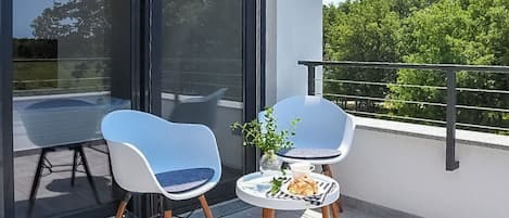 Un balcón con dos sillas, una mesita con una planta y una taza, y una puerta de vidrio que da a un espacio al aire libre.