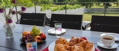 A dining table with croissants, fruit, and coffee, set on an outdoor terrace with chairs and a view of greenery.
