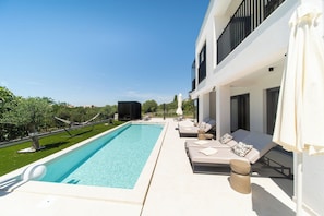 Un área moderna junto a la piscina con una pileta rectangular, sillas de descanso y un edificio blanco con balcones.