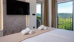 A hotel room with a bed, a flat-screen TV mounted on the wall, and a balcony with a view of greenery. - Villa Loparia (Lopar)