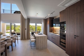 A modern kitchen with a central island, bar stools, and a dining area with a view of greenery.