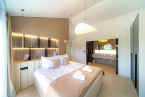 A modern hotel room with a large bed, bedside tables, a built-in desk, a large window, and a bathroom with a mirror and sink.