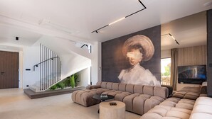 A modern living room with a curved sectional sofa, a round coffee table, and a staircase with a unique design. - Villa Loparia (Lopar)