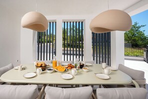 A dining table set with breakfast items, two hanging pendant lights, and a view through a window with vertical blinds. - Villa Loparia (Lopar)