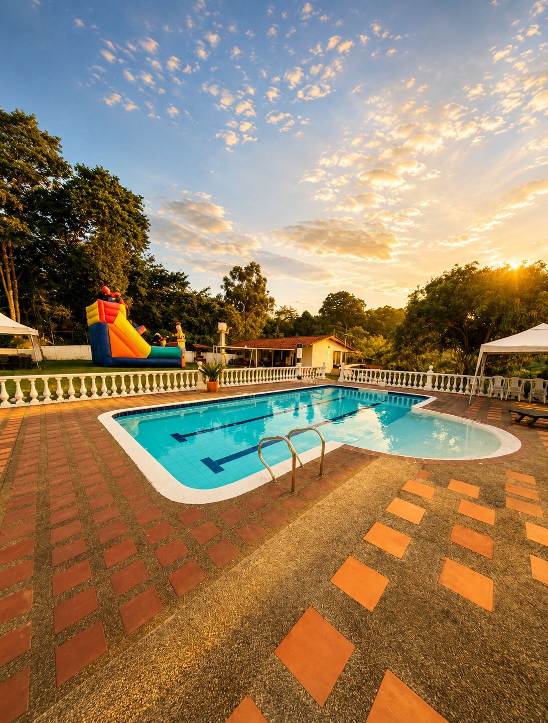 Une piscine avec un toboggan et des jeux gonflables, entourée d’une terrasse en briques et d’une clôture blanche.