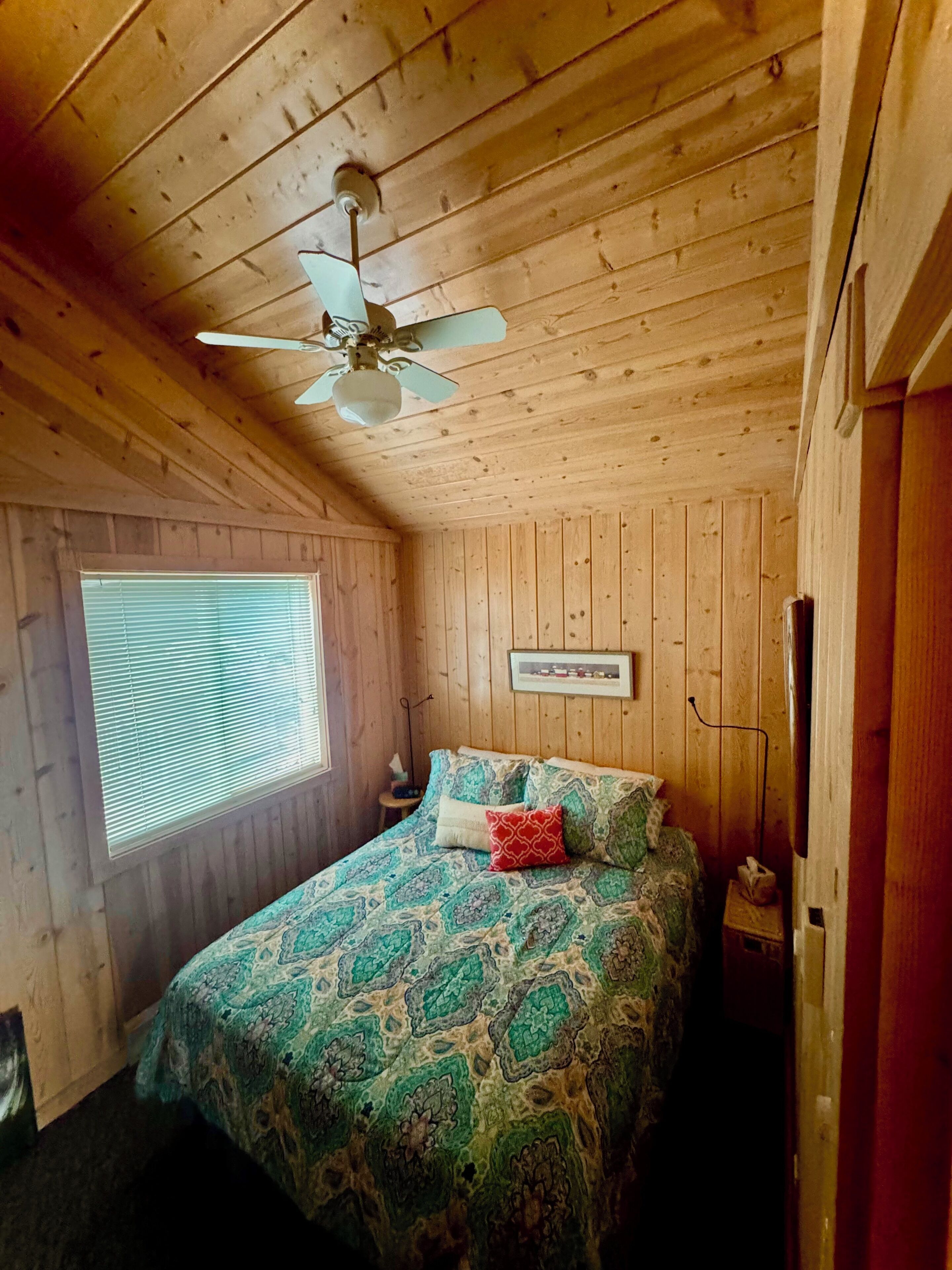 A bedroom with a wooden ceiling, a bed with patterned bedding, a ceiling fan, a window with blinds, and a framed picture on the wall.