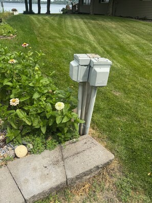 A utility box next to a flower bed with green foliage and a clear view of a lake in the background. - Cozy lake cabin with 50 Amp for RVs on Pelican Lake, with private dock. (Detroit Lakes)