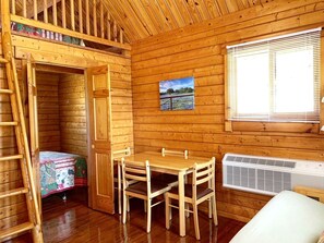 A wooden cabin interior with a dining table, chairs, a ladder, a bed, and a window with blinds. - Peaceful Lakefront Cabin with Loft, Beach Access & Sunset Views in Buchanan Dam, Texas (Buchanan Dam)