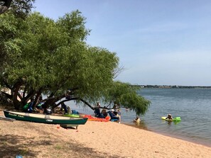 A beach with a row of boats, a tree, and people relaxing by the water. - Peaceful Lakefront Cabin with Loft, Beach Access & Sunset Views in Buchanan Dam, Texas (Buchanan Dam)