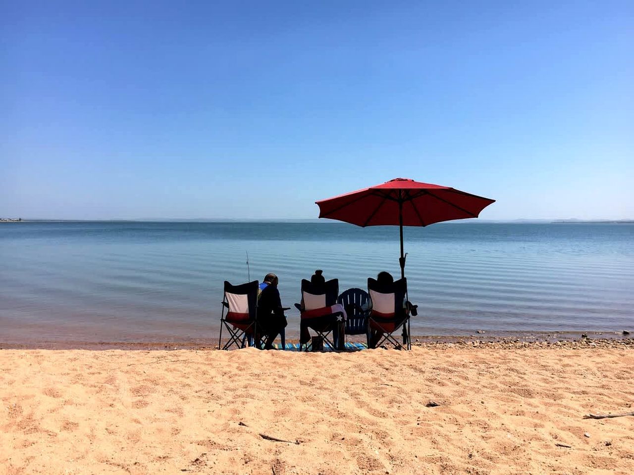 A beach with three chairs under a red umbrella, a calm sea, and a clear sky.