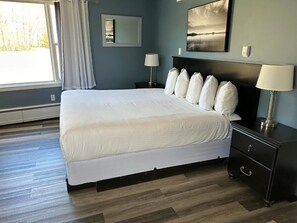 Un dormitorio con una cama grande, dos mesitas de noche con lámparas, un cabecero con varios cojines, un espejo fijado a la pared y un cuadro enmarcado.