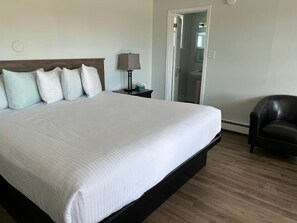 Habitación de hotel con una cama grande, una mesita de noche con lámpara, una silla y un baño visible a través de una puerta abierta.