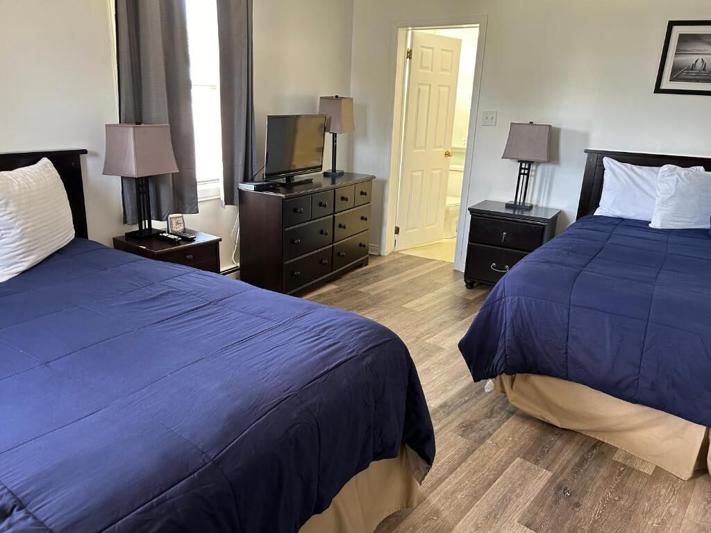 Quarto de hotel com duas camas, uma cômoda, uma televisão e uma porta que dá para outro quarto.