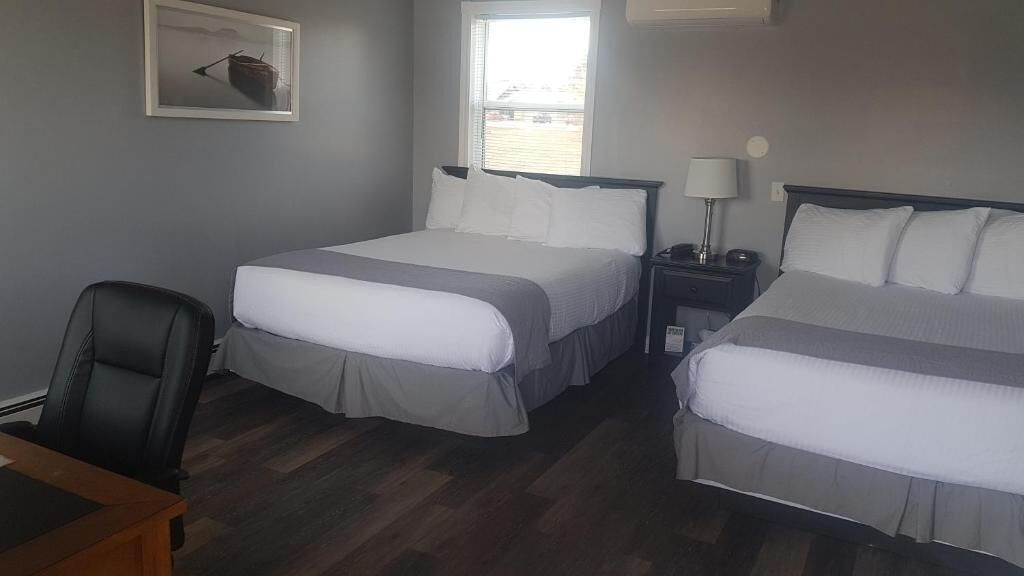 A hotel room with two beds, a desk, a chair, a nightstand with a lamp, and a window with blinds.