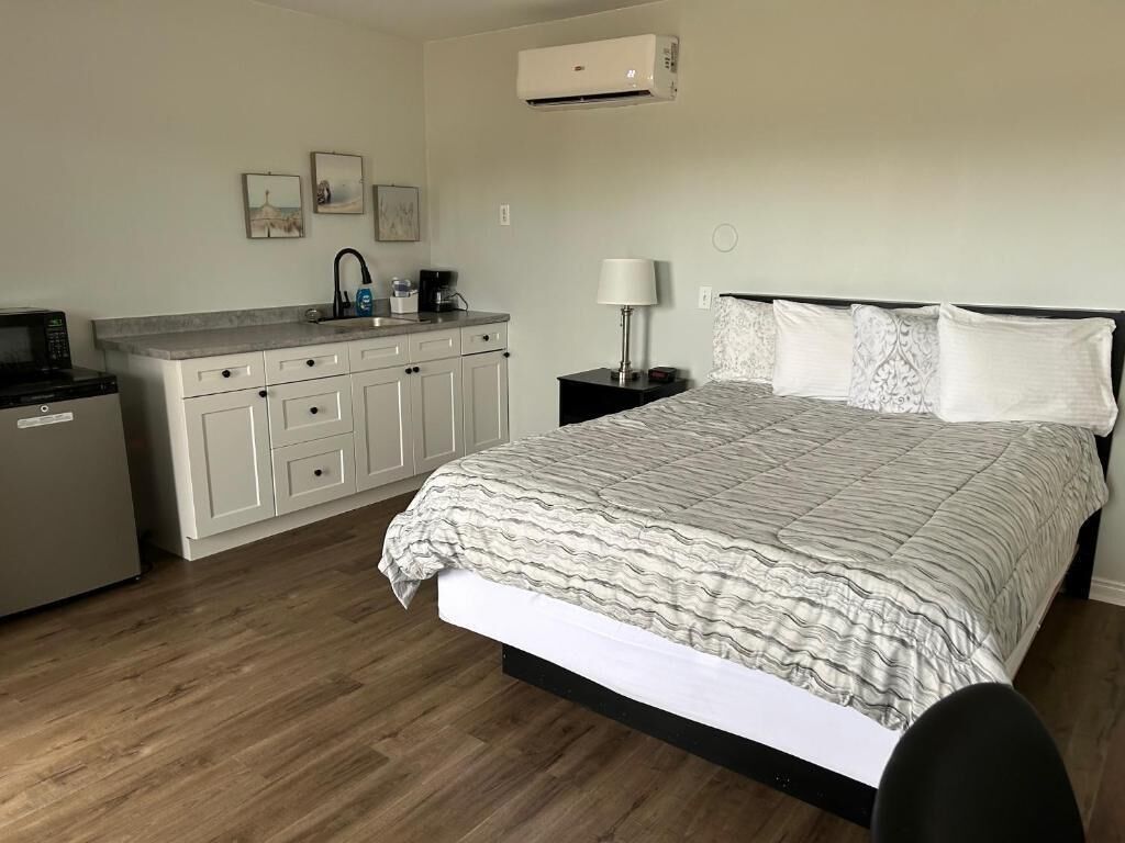 A bedroom with a bed, a nightstand, a lamp, a desk with a computer, and a kitchenette.