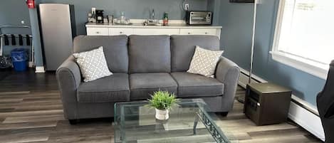 A living room with a sofa, a glass coffee table, a kitchen area with a microwave, and a window.
