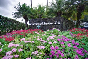 A stone wall with a sign reading 'Regency at Poipu Kai' surrounded by a garden with pink and red flowers. - Regency at Poipu Kai 823 | Spacious 3BR Townhome Near Poipu Beach (Koloa)