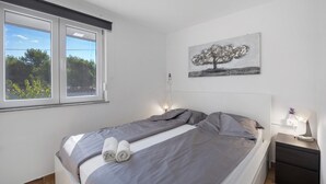 Quarto com cama, mesa de cabeceira, abajur e janela com vista para o verde.