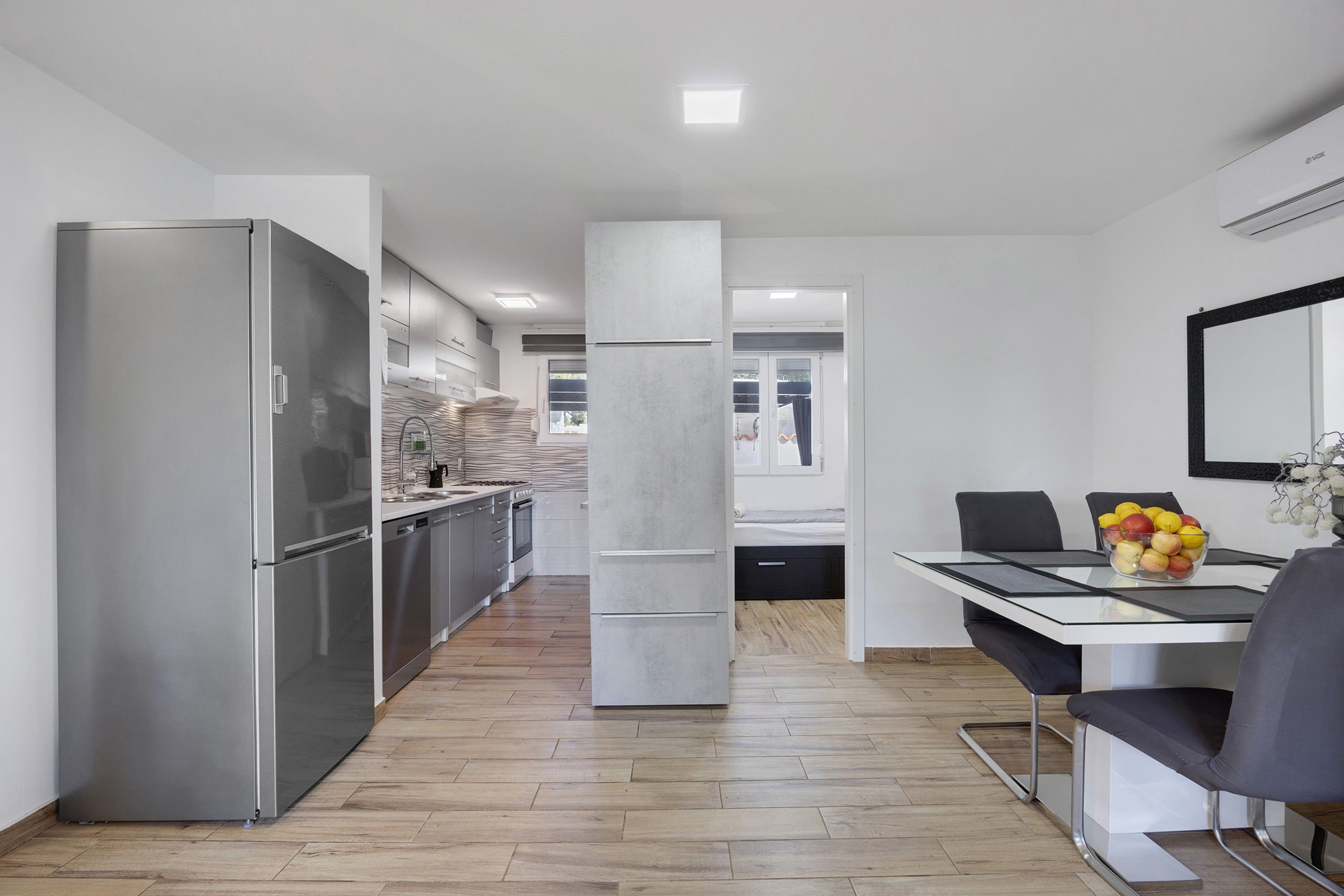 Een moderne keuken met roestvrijstalen apparatuur, een eethoek met een glazen tafel en uitzicht op een slaapkamer.