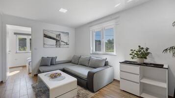 Ein modernes Wohnzimmer mit einem grauen Ecksofa, einem weißen Couchtisch, einem kleinen weißen Schrank und einer Topfpflanze.