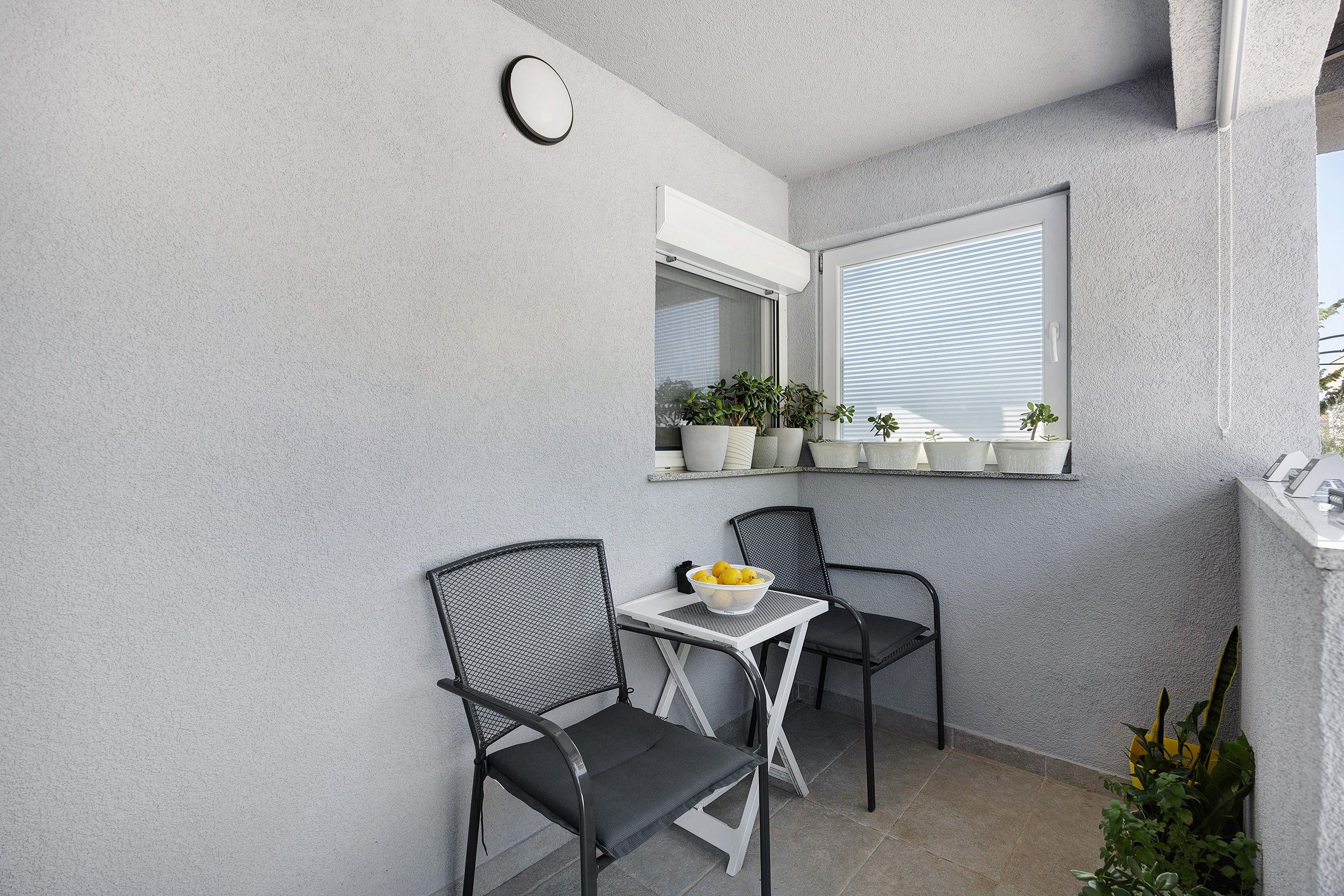 Un pequeño espacio al aire libre con dos sillas, una mesa, una ventana con persianas y un arreglo de plantas en macetas en la pared.