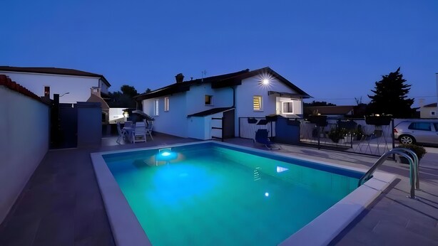 Piscina residencial com uma casa ao fundo, cercada por uma vedação e árvores.