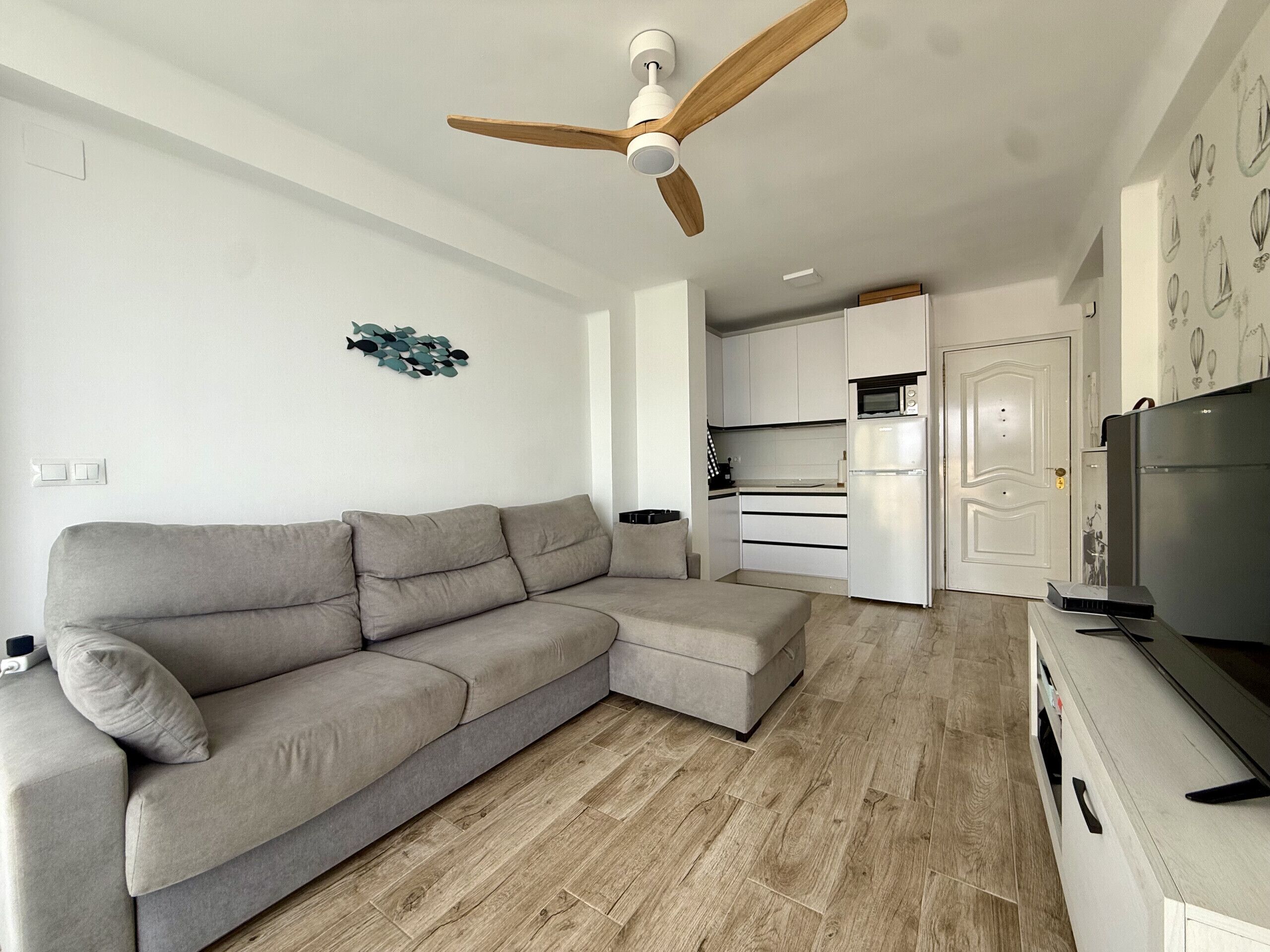 A modern living room with a grey sectional sofa, a ceiling fan, a kitchen area with white cabinets, and a flat-screen TV.