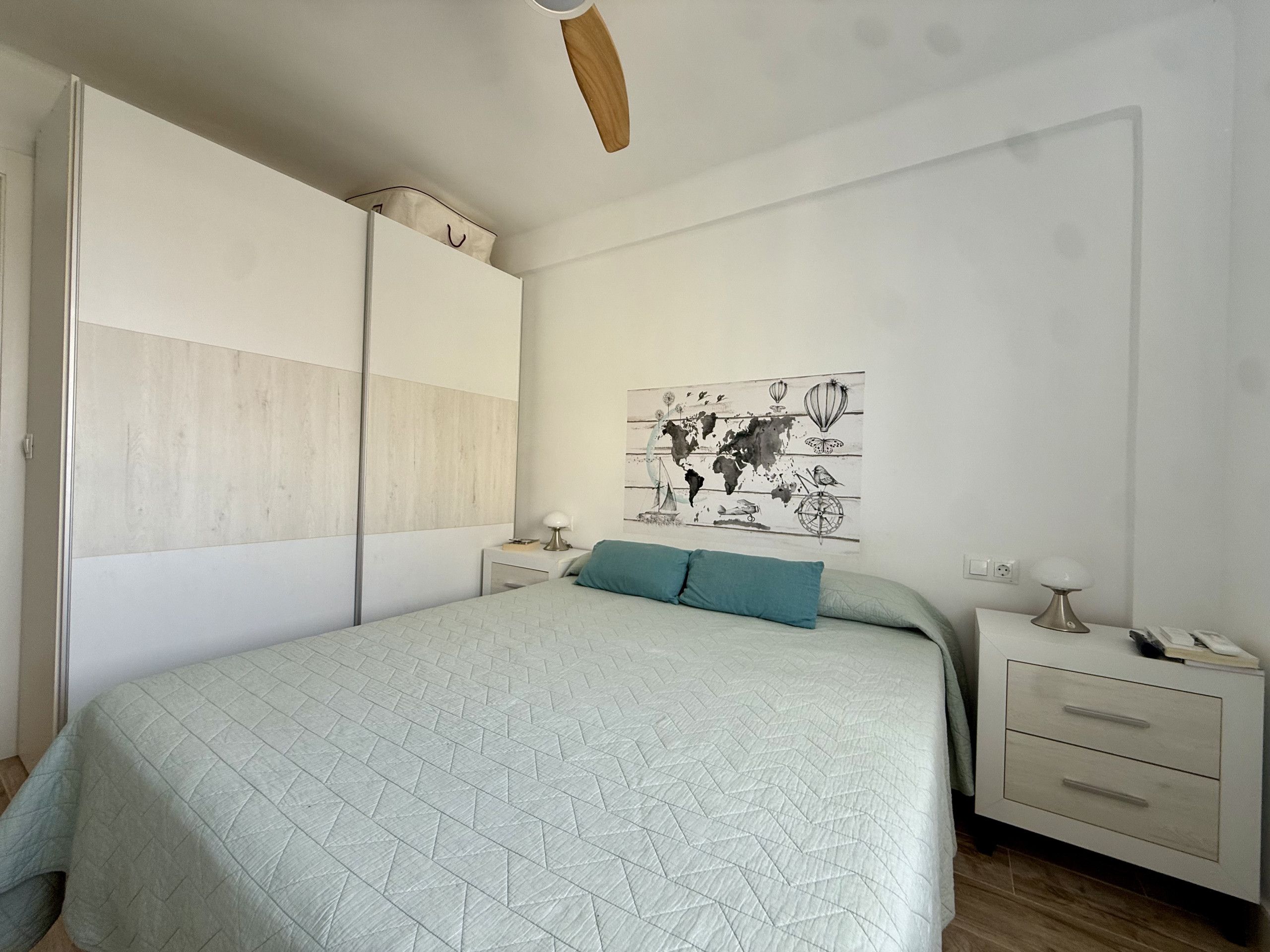 Une chambre à coucher comprenant un lit, une table de chevet, une commode, une œuvre d’art représentant une carte du monde et un ventilateur de plafond.