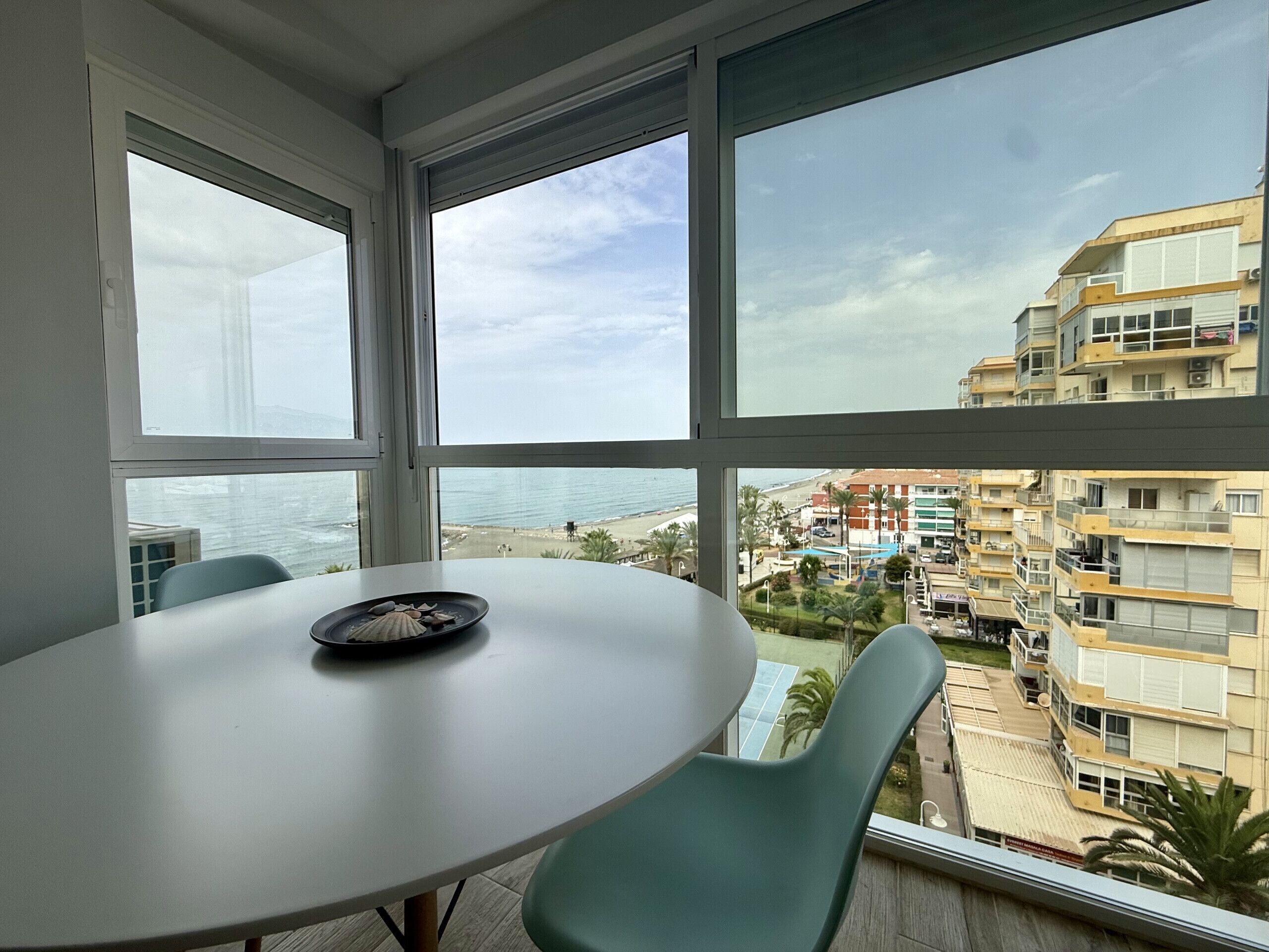 A room with a round table and two chairs, a window with a view of the beach and buildings.