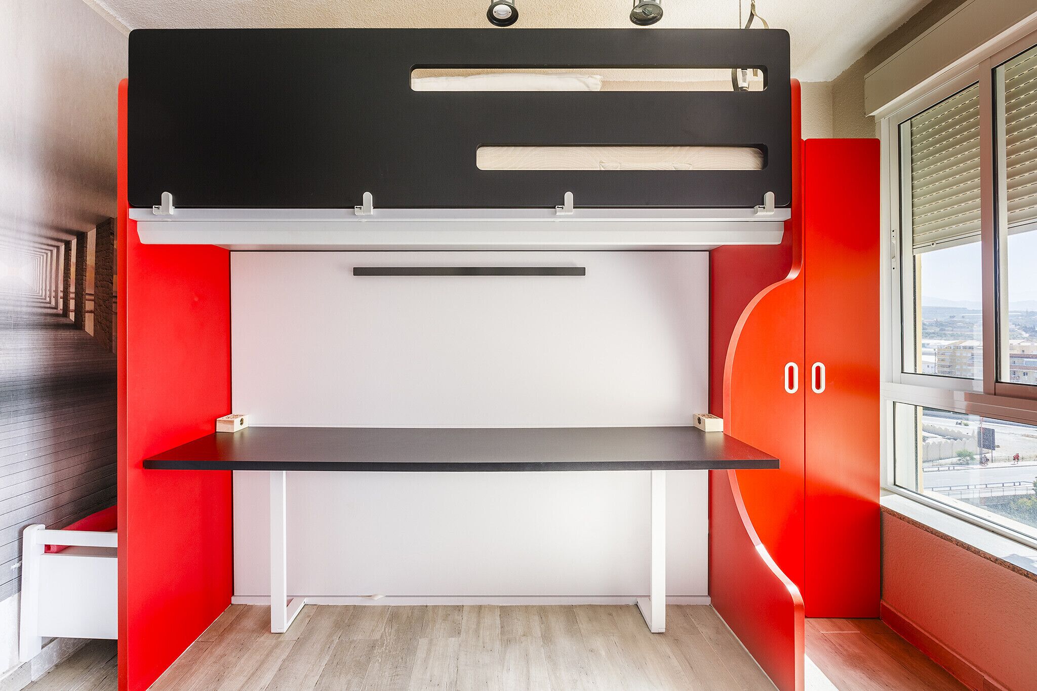 A bunk bed with a desk in the middle, a window with blinds, and a view of the city.