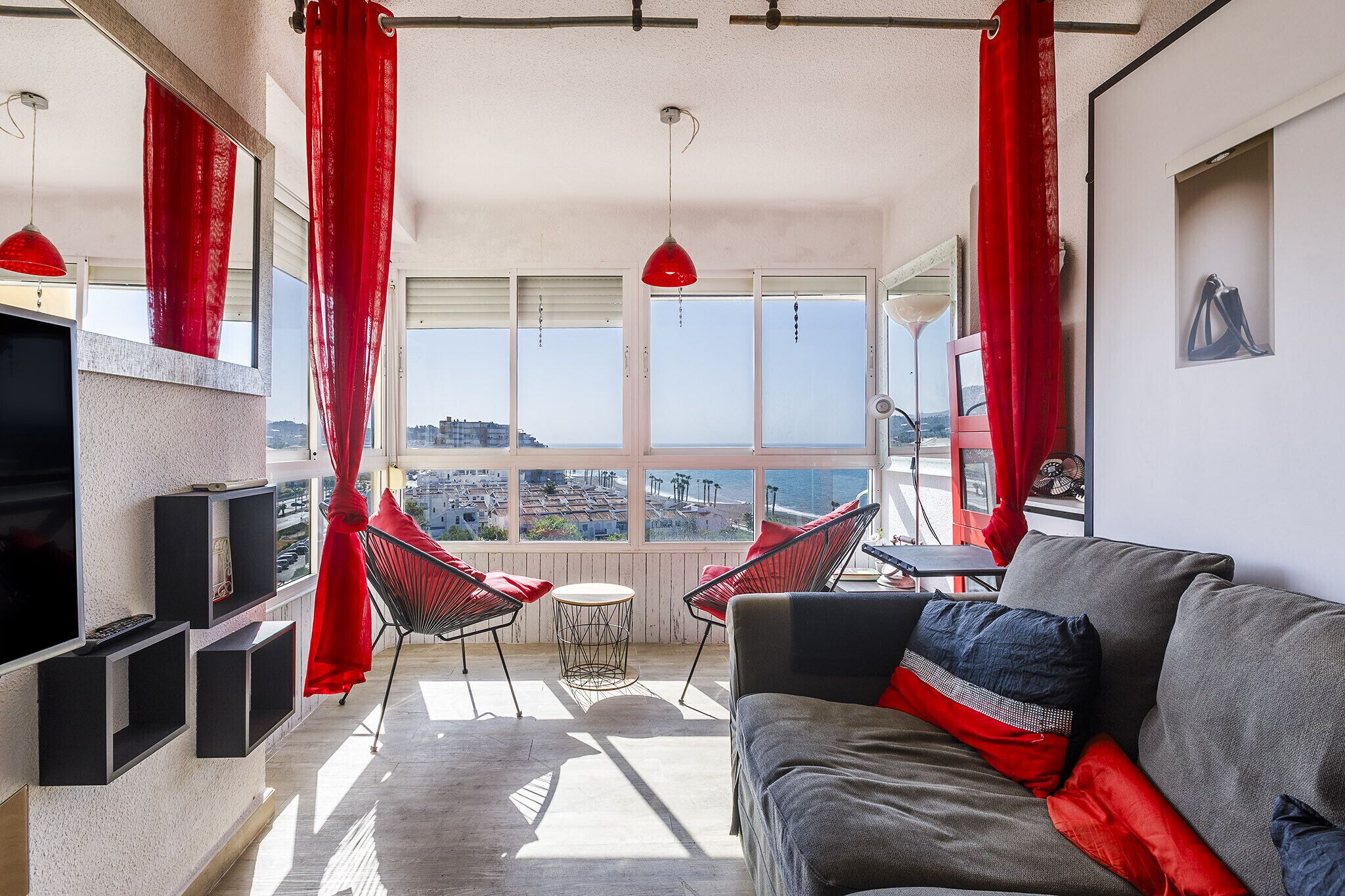 Ein modernes Wohnzimmer mit grauem Sofa, roten Vorhängen und Blick auf die Stadtlandschaft.
