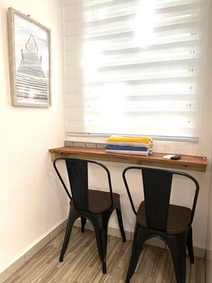A room with two black chairs, a wooden shelf with towels, a framed picture, and a window with blinds. - Stay Near Everything & Beach - Walkable Condado (San Juan)