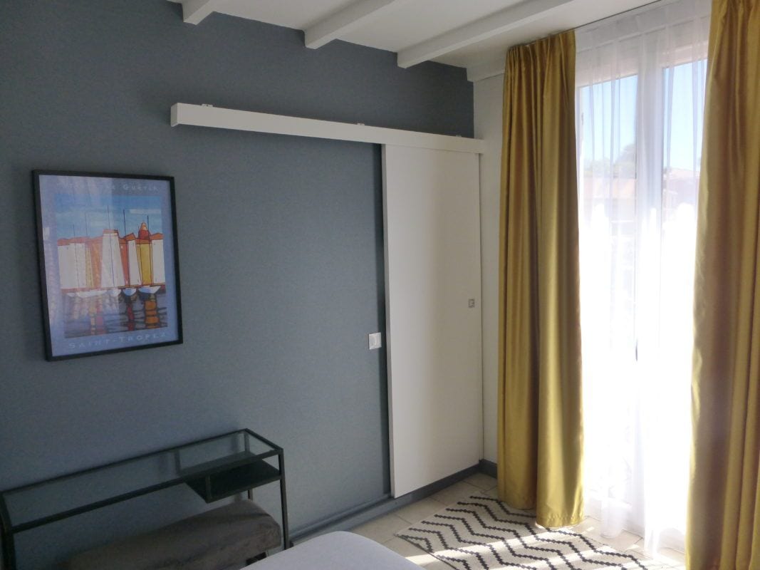 Ett hotellrum med en säng, ett sängbord, en inramad tavla på väggen, en garderob och ett fönster med gardiner.