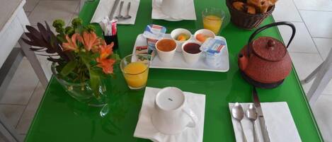 Una mesa de desayuno con una bandeja verde, una tetera, una cesta de croissants y un florero con flores.